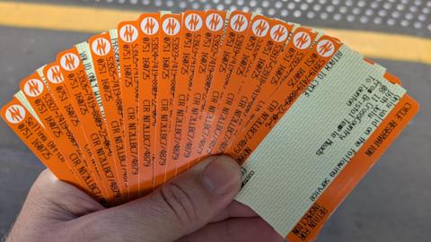 A man standing on a train platform holding a large fan of 15 orange train tickets in his left hand. 