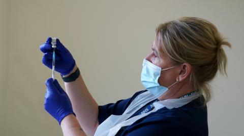 A blonde nurse, wearing a face mask, is preparing a vaccination.