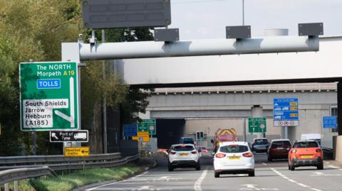 The entrance to the Tyne Tunnel. Several cars are driving towards it. There is a green road sign to the left of the road, it says 'TOLLS'. The exterior of the tunnel is a mixture of cream and grey brick, with cream bricks making up the top half, and grey bricks the bottom. There are also green trees to the left of the road.