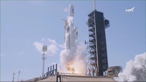 Image shows a white rocket launching from the launchpad, with white smoke visible at the bottom and an orange fire