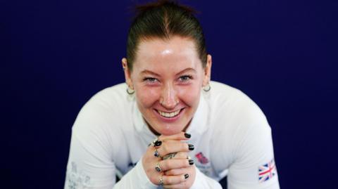 Makayla Gerken Schofield pictured in a Team GB publicity shoot. She is wearing a white long-sleeved top and is leaning on their hands. She has dark hair pulled back and is smiling.