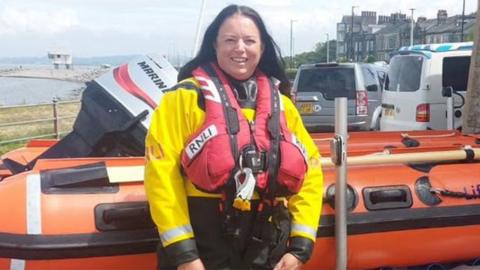 Fiona, who had just completed a year's training, who has long black hair, standing by n orange lifeboat wearing a yellow jacket and red life vest