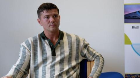 A young man with short dark hair is sitting and looking to the right of the picture. He is wearing a long-sleeved top comprising wide, vertical grey and white stripes. 