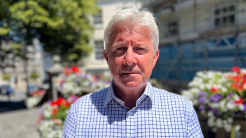 A grey-haired man in a blue and white checked shirt stands in front of a public building, with flowers and trees behind him.