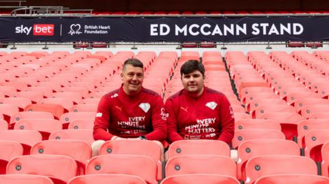 Two men in red shirts sit among red stadium seats. A banner behind them reads Ed McCann Stand