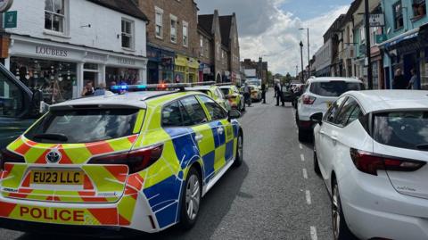 A line of police cars in a street with shops and restaurants on either side of the road. Police officers and pedestrians are on the road and pavement. 