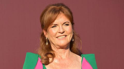 Sarah Ferguson stood in front of a dark red back drop. She is wearing a green dress with pink parts to it. 