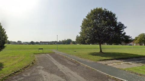 A  stretch of green playing field in Foxbridge with a few trees and houses in the distance.
