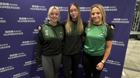The three ladies stand in rugby tops in front of a BBC Radio Humberside screen.