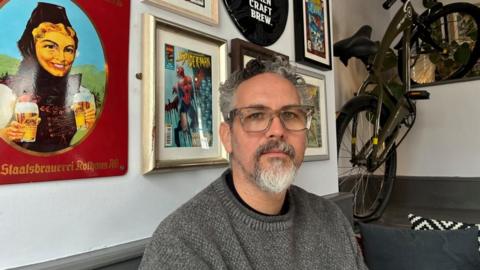 Sam Cornwall-Jones co-owner of the Magic Wingdom - sat in the Drawing Board pub with images including comic book posters on the wall and a bike mounted on the wall behind him.
