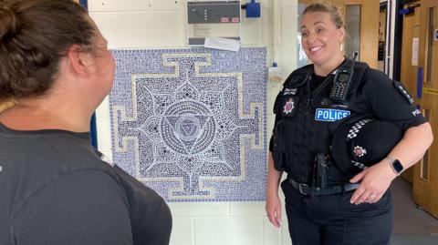 Tendring district commander Chief Inspector Ella Latham in police uniform, smiling and looking at another woman while standing by a blue mosaic on a wall