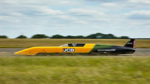 A yellow and black car driving on a track. There is grass in the foreground, and the background is blurred by the speed of the car.