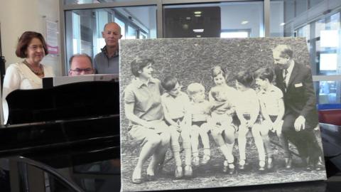 Christopher sat behind a piano and Jeremy and Susan behind him as he plays. In the front of the image is their black and white family photo.