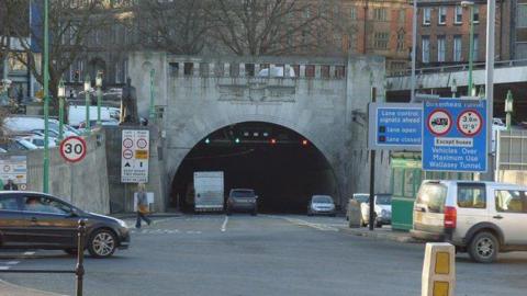 The arched, grey entrance to the Queensway Tunnel, with cars driving into the darkness