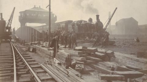 A brown and white photo of a train track recently constructed with piles of rectangular panels in the middle. A train can be seen going under a bridge in the distance with steam coming from it. Several men are stood around the tracks.