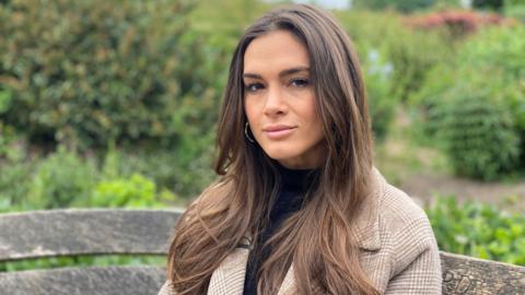 A woman with long brown hair and is wearing a light brown and white coat. She is sitting at a garden. 