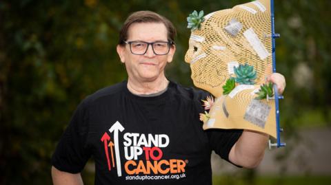 A man with brown hair and glasses wearing a black t-shirt with a Stand Up To Cancer logo in the middle holds up a face shaped object made of a flesh coloured mesh that is covered in small plants.