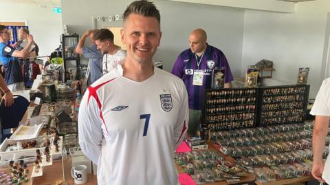 Adam Grocott smiles at the camera, wearing a white Umbro long sleeved England shirt with a red cross on the right shoulder and a blue number 7 in the middle. To his left is a table with hundreds of Corinthians models on, and more to his right. Other tables with memorabilia are behind and people manning the stand or milling about.
