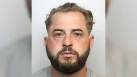 A police custody photo of Ashley Sherred. He has brown hair, brown eyes, a dark-brown beard, and is wearing a grey top.