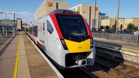 A Greater Anglia train in white, red and grey livery waits at a platform. It's a clear, sunny day.