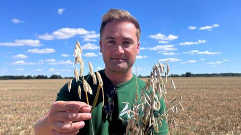 Tristan Ashby, standing in a field, holding onto some crops. A field is behind him with crops in it. He is wearing a green T-shirt, with sunglasses hooked on the front. He has short blonde hair. 