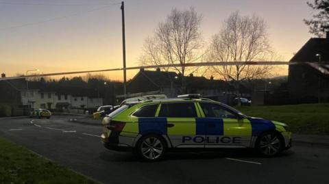 A yellow blue and white police car sits in the middle of the road at a security alert in Twinbrook