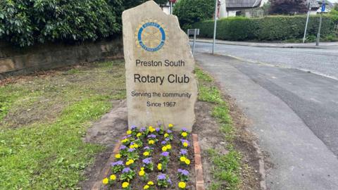 The newly installed stone marker on Liverpool Road, Penwortham, which reads Preston South Rotary Club - Serving the community since 1967. Purple and yellow flowers are planted at the foot of the stone marker.