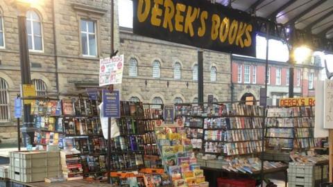 Derek's Books stands on the edge of Preston outdoor market. The stall has lots of books neatly stacked on stands.