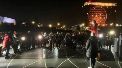 Group of people in warm winter clothes stood in the dark with phone lights listening to a male speaker.
