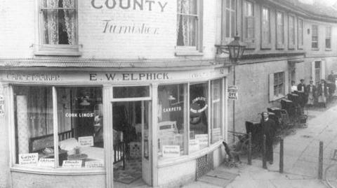 A old black and white photo of the original Elphicks on the High Street. It has a round front and the letters 'E.W Elphick' painted above the door. There are signs in the shop windows for dye, carpets and cork linos. 