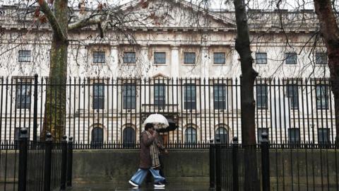 A woman holding an umbrella past black railings in front of the Royal Mint Court, the proposed site of the new Chinese embassy in London.