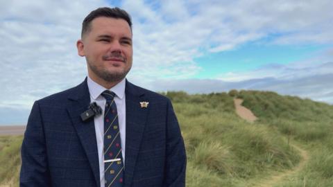 Lancashire County Councillor Joshua Roberts on the sand dunes at Lytham St Annes. The Reform UK politician is wearing a dark blue suit and tie. He has dark hair and is smiling.
