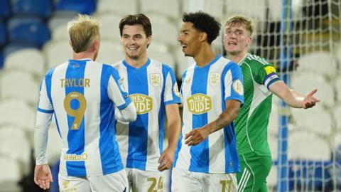 Huddersfield town players Joe Taylor, Leo Castledine and Ruben Roosken talk and smile with each other wearing blue and white vertical stripe shirts while newcastle under 21s captain Cathal Heffernan holds an arm out wide with a protesting look on his face 