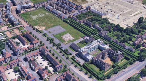 An aerial picture. To the left is a section of new build housing. In the middle is an empty green area with a car park. To the right of it is an older style building. A tree lined street is beyond that with a flat construction area further afield. 