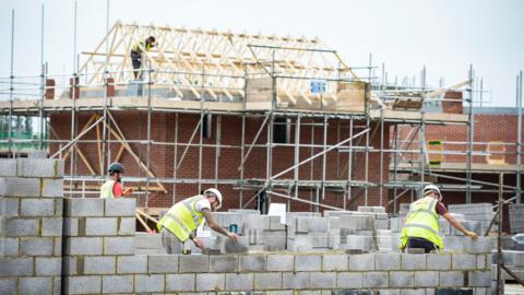 Builders placing bricks onto a wall with a house with scaffolding in the background.