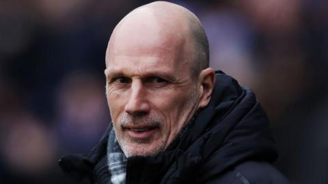 Philippe Clement has a grey stubble, shaved head and is wearing a black coat and checked scarf. He is slightly smiling with his head turned towards the camera.