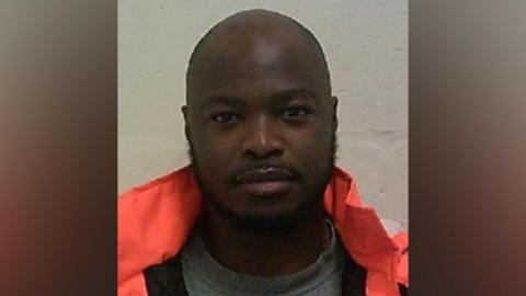 A bald man with a beard and moustache in a police mugshot. He is wearing a bright orange coat.