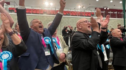 Reform UK members celebrate during the local election count for Plymouth City Council. They are mostly wearing suits and have Reform rosettes pinned to their jackets.