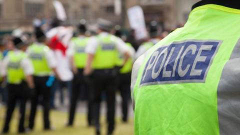 a close-up of a police vest in the foreground, as a blurred group of people with placards and a group of police officers are gathered outside a building in the background