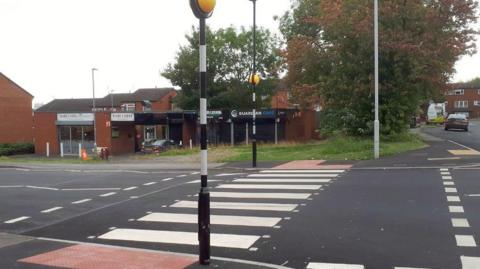 A zebra crossing that is raised slightly by a junction. A single-storey building can be seen on the far side of the road, and residential properties.