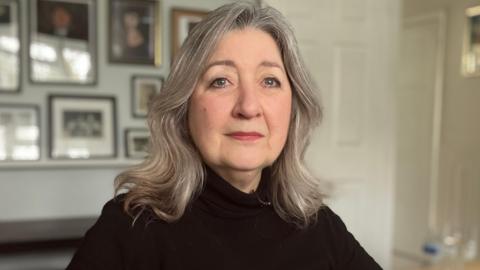 Diane Cleveland is seen from the shoulders up facing towards us. She is sitting in her living room wearing a black jumper and has shoulder-length hair. Pictures can be seen on the wall behind her