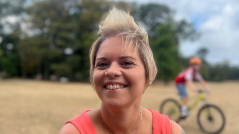 A woman with dyed blonde hair smiles at the camera, wearing a red dress. In the background is a blurred field and a child on a bike.