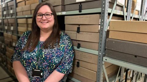 A woman in a floral top with short sleeves stands in an archive room with cardboard boxes stacked on shelves behind her. She is wearing a lanyard, glasses and has long brown hair.
