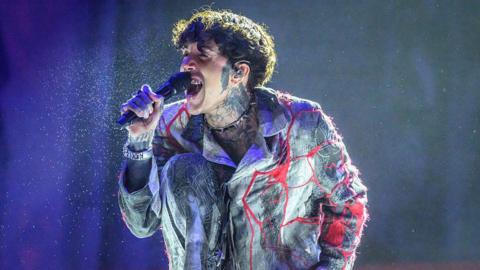 Oli Sykes performing onstage during Leeds Festival at Bramham Park in 2025. He has curly brown hair. He is singing into a mic wearing a red and grey shirt-jacket. He has tattoos on his hands wrists, neck and face. 