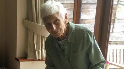 Judy Zussman, with short grey hair and wearing a black and white top with a mint green coat, is photographed standing in front of a window with cream tied-back curtains.