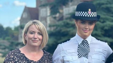 A woman with shoulder-length blonde hair standing next to police officer Hannah Byrne, who is wearing her police uniform and hat.