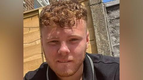 A young man with short strawberry blonde hair and a nose piercing wearing a black polo shirt