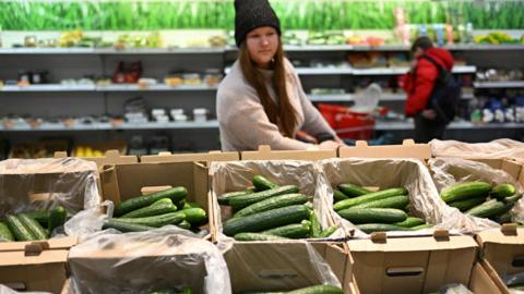 A woman with long dark hair wears a black hat while walking through a supermarket. She looks at the selection of cucumbers on offer.