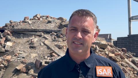 Lang stands in front of a pile of bricks, concrete and rubble He wears a dark top with UK Fire and Rescue ISAR orange badge.