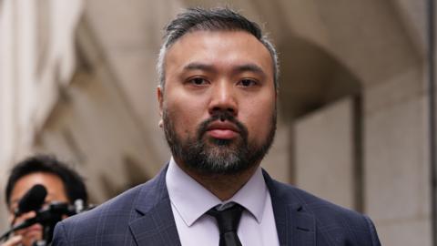 A man looks into the camera outside of a court, wearing a formal suit. A photographer can be seen behind him out of focus.